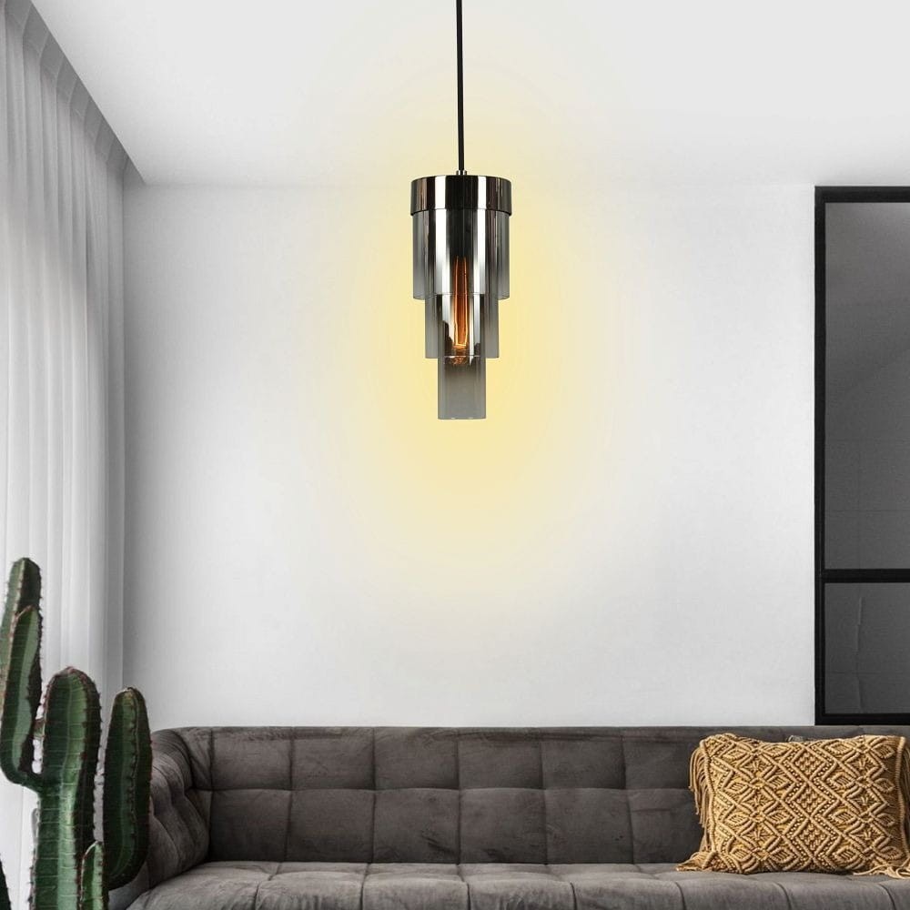 Lampa wisząca Grans 1xE27 w kolorze czarnym i chromowanym, o cylindrycznym kształcie, zawieszona na cienkim przewodzie, emitująca ciepłe światło. W tle widoczny szary narożnik sofy oraz roślina doniczkowa.