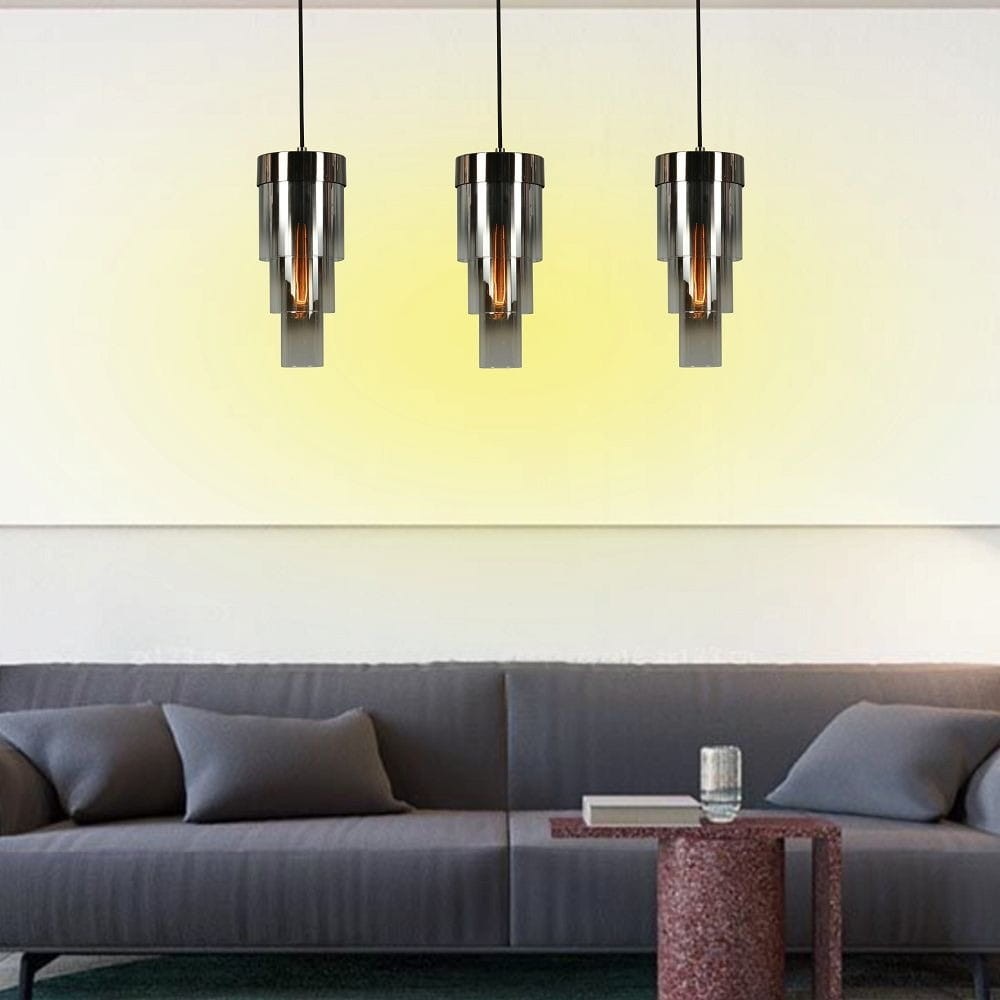 Lampa wisząca Grans 3xE27 w kolorze czarnym z chromowanymi detalami, zawieszona na długich przewodach. Posiada cylindryczny kształt, z widocznymi żarówkami wewnętrznymi, umieszczona nad szarym sofą w nowoczesnym wnętrzu. Tło w jasnym kolorze.