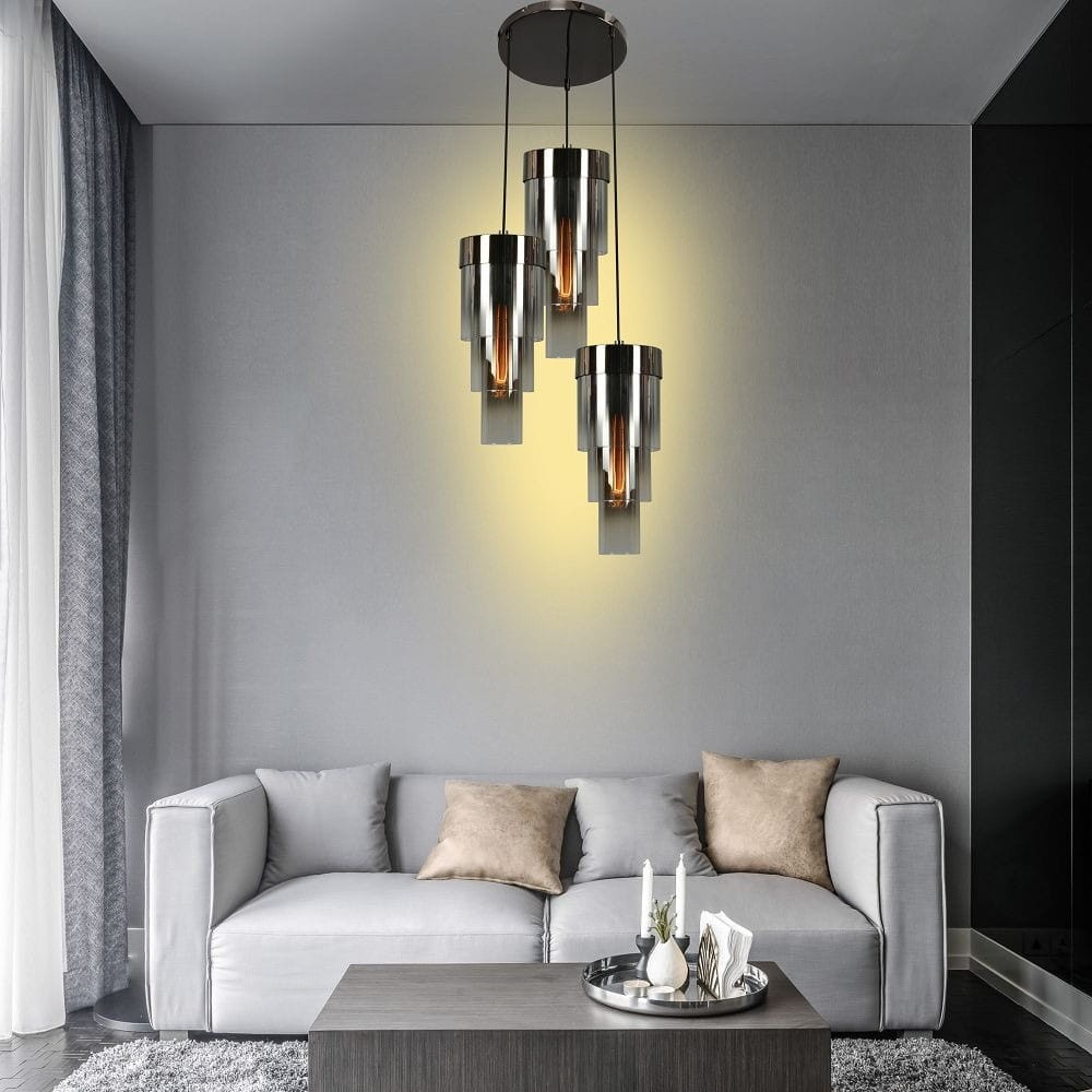 Lampa wisząca Grans 3xE27 w kolorze czarnym i chromowanym z trzema cylindrycznymi kloszami. Zawieszona na długich linkach, emitująca ciepłe światło na tle szarej ściany, w aranżacji salonu z jasną sofą i drewnianym stolikiem.