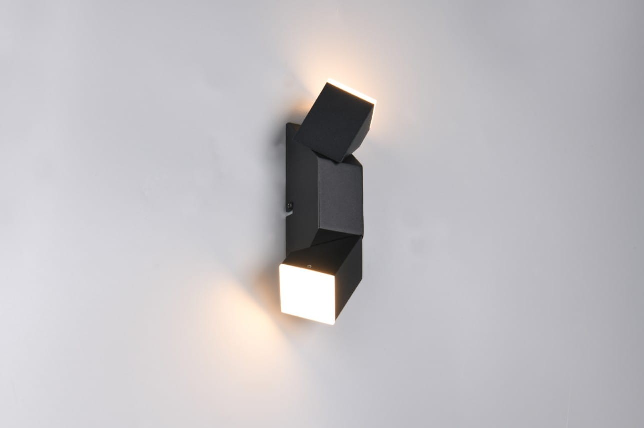 Raglan kinkiet w kolorze czarnym z dwiema głowicami LED, oświetlającymi przestrzeń. Produkt zamontowany na ścianie, w nowoczesnym, kształcie geometrycznym. Widoczna jedna matowa, czarna płaszczyzna oraz jedna biała, emitująca światło.