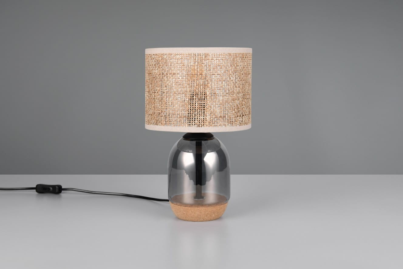 Lampa stołowa Tara z kloszem z naturalnego materiału i podstawą z przezroczystego szkła oraz korka. Ujęcie frontalne, lampa stoi na płaskiej powierzchni. Kolor chromowany podstawy i beżowy klosz w stylu minimalistycznym.