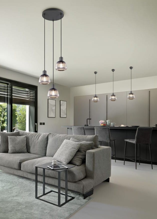 Lampa wisząca Tarifa 3xE27 w kolorze czarnym z trzema szklanymi kloszami, zawieszona na długich linkach. W tle nowoczesna kuchnia z ciemnymi meblami i szarym narożnikiem. Proste, eleganckie wzornictwo.