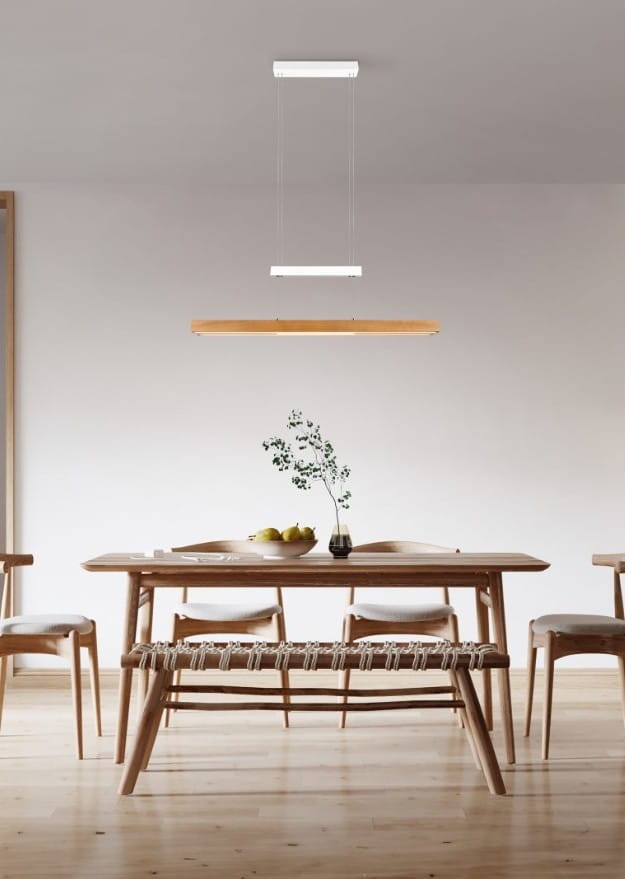 Lampa wisząca Trajan z trzema źródłami LED, w białej obudowie i jasnodrewnianym kloszu. Zawieszona nad drewnianym stołem w nowoczesnej jadalni, w otoczeniu krzeseł i dekoracyjnej rośliny w wazonie.