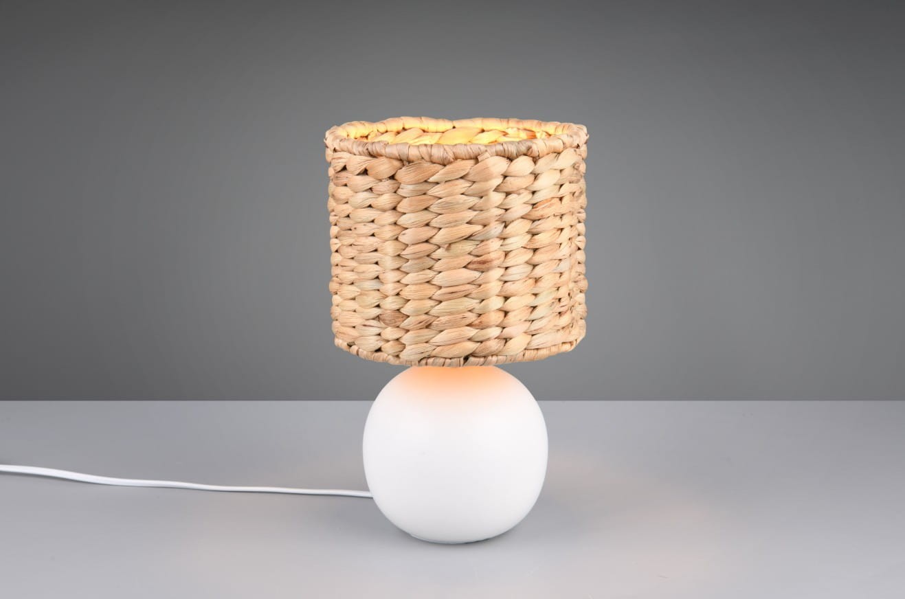 Lampa stołowa Vina z białą, okrągłą podstawą i kloszem z plecionki w jasnym kolorze. Ujęcie frontalne, lampa stoi na gładkiej powierzchni. Widoczny kabel zasilający. Klosz ma otwarte wykończenie, co umożliwia rozprzestrzenianie światła.
