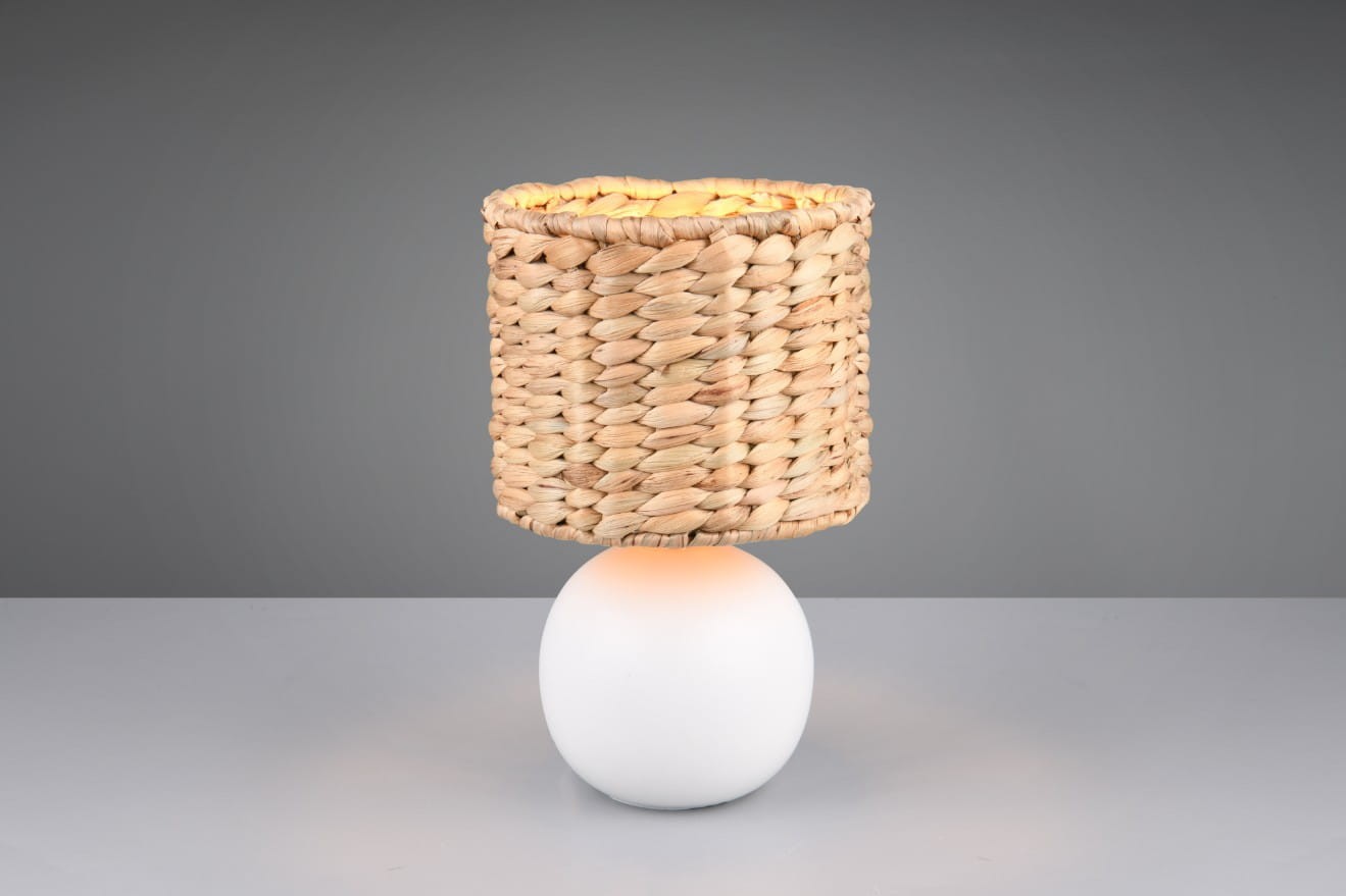 Lampa stołowa Vina z białym, okrągłym podstawą i kloszem z plecionki w odcieniach naturalnych. Ujęcie z boku, ukazujące detal struktury klosza. Wyposażona w jeden gwint E27, idealna do nowoczesnych aranżacji.