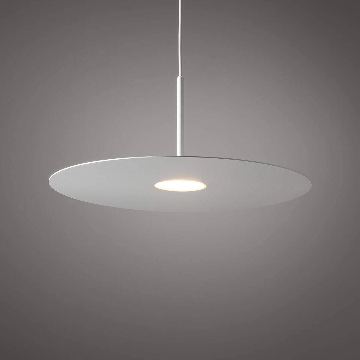 Lampa wisząca Simone 1xGX53 w kolorze białym, o okrągłym, płaskim kloszu zawieszonym na cienkim, metalowym pręcie. Źródło światła umieszczone w centralnej części klosza. Tło w odcieniu szarym, minimalistyczny design.