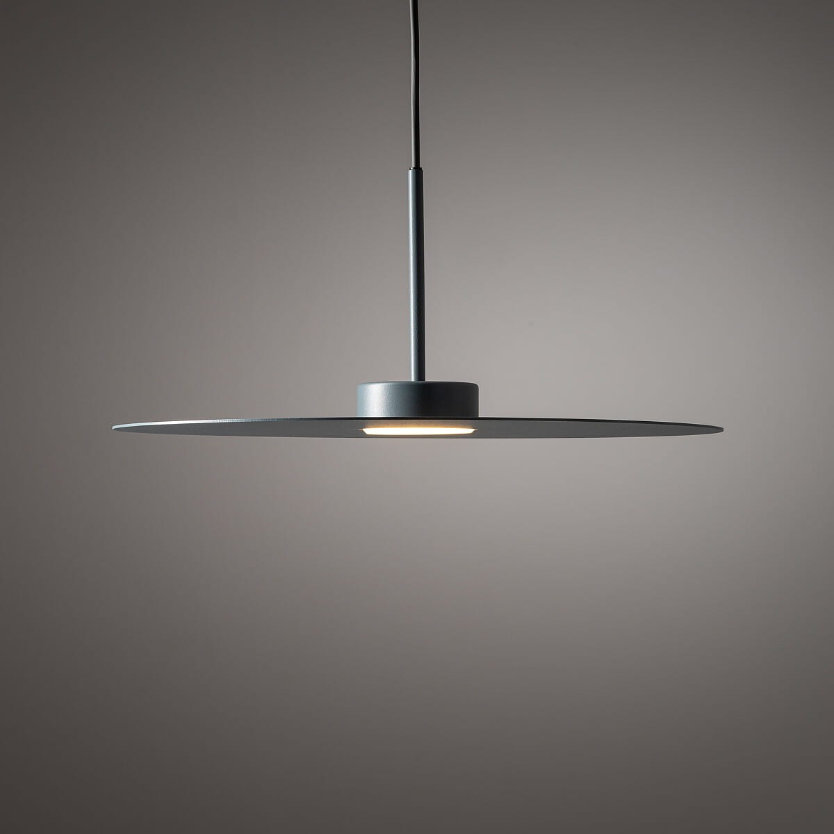 Lampa wisząca Simone w kolorze niebieskim z pojedynczą oprawą GX53. Posiada okrągły, płaski klosz zawieszony na cienkim przewodzie. Umieszczona centralnie, emituje światło w dół. Tło w odcieniu szarym, minimalistyczne.
