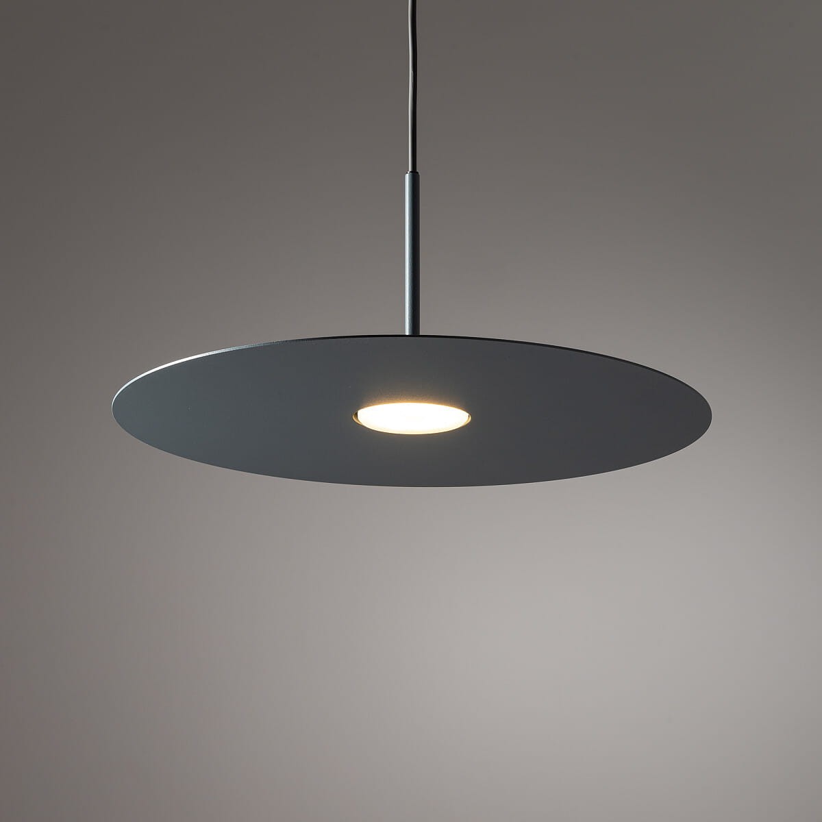 Niebieska lampa wisząca Simone z jednym źródłem światła GX53, w kształcie okrągłej, płaskiej tarczy, zawieszona na cienkim przewodzie. Elementy metalowe w matowym wykończeniu, w centralnej części widoczna żarówka. Tło w odcieniach szarości.