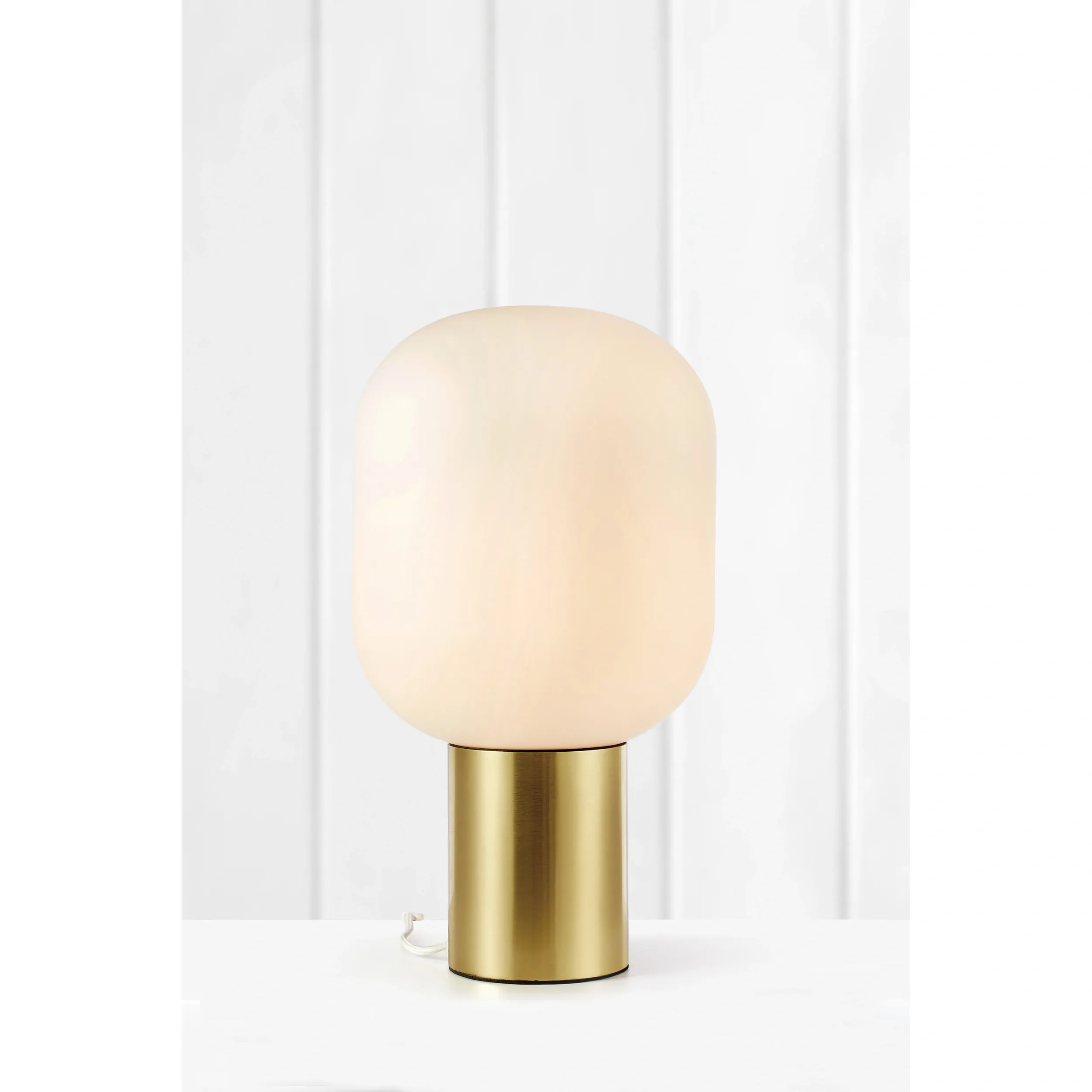 Lampa stołowa Brooklyn 1-punktowa w kolorze złotym z białym kloszem. Okrągły, matowy klosz na cylindrycznej podstawie. Ujęcie frontalne, na jednolitym tle. Widoczne przewody zasilające. Wysoka, nowoczesna forma.