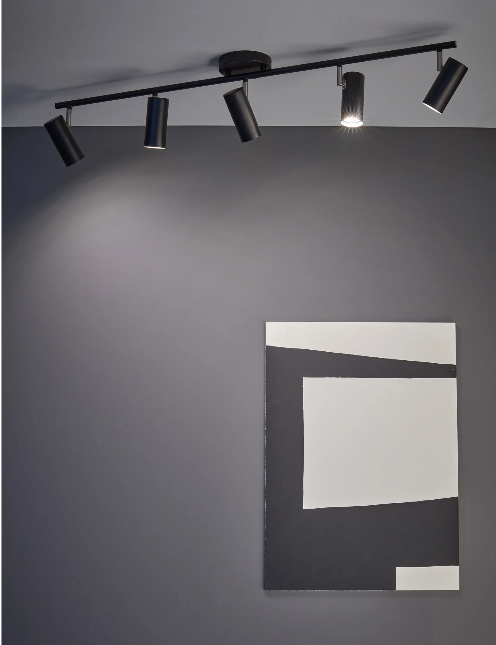 Czarna lampa sufitowa 5-punktowa Barcelona z metalowymi reflektorami w kształcie cylindrów, zamontowana na prostokątnej podstawie. Ujęcie z góry, w tle szara ściana i nowoczesny obraz w odcieniach bieli i czerni.
