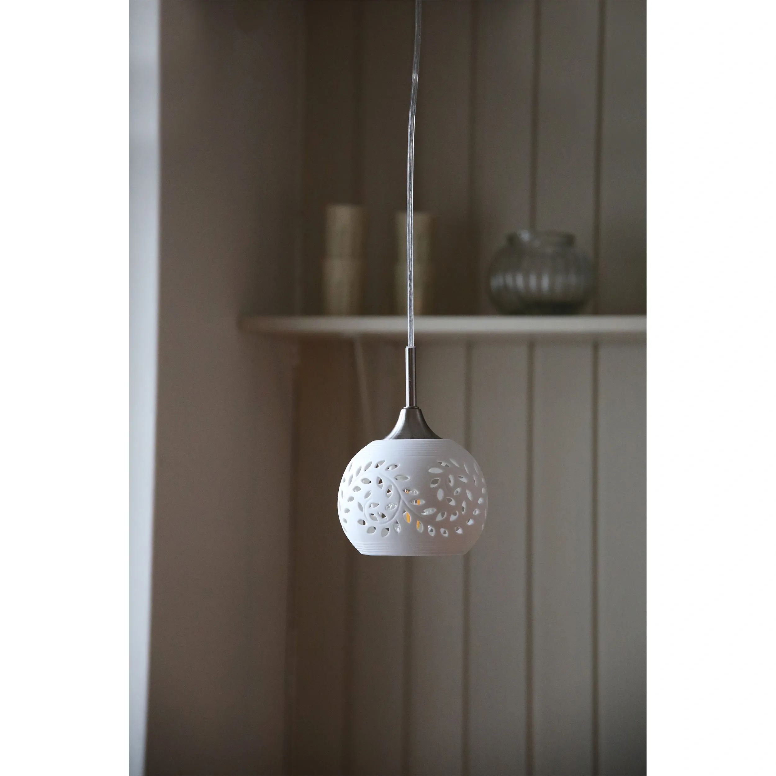 Lampa wisząca Lohals w kolorze białym z metalowym elementem, o okrągłym kształcie z wyciętymi wzorami. Zawieszona na cienkim przewodzie, z widocznym detalem w postaci dekoracyjnych otworów, wisi na tle jasnej ściany.