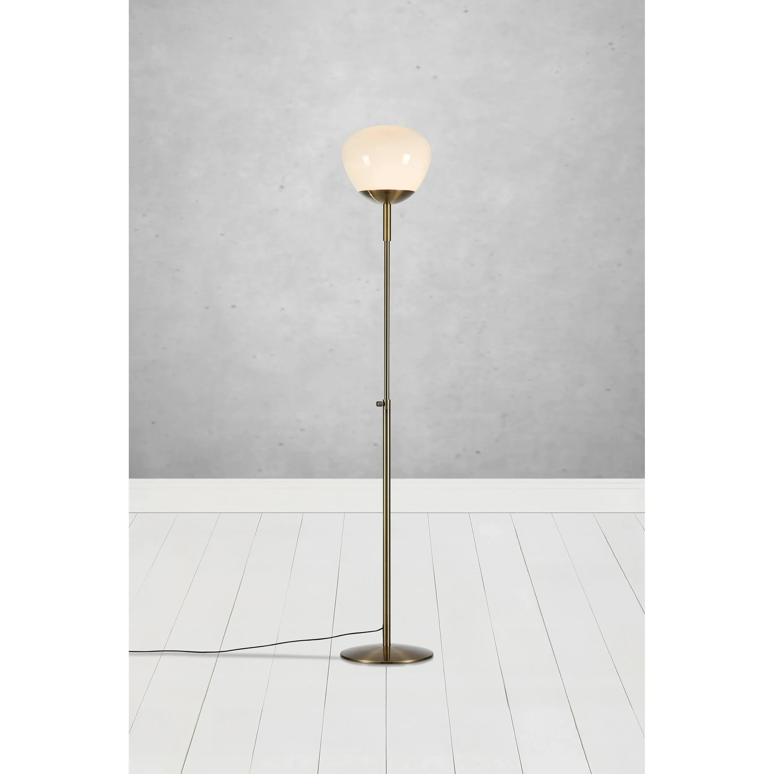 Lampa podłogowa Rise w kolorze mosiądzu z białym kloszem. Stojąca konstrukcja z metalowym stelażem, z widocznym kablem. Klosz o zaokrąglonym kształcie umieszczony na wysokości około 150 cm, na tle szarej ściany i białej podłogi.