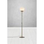 Lampa podłogowa Rise w kolorze mosiądzu z białym kloszem. Stojąca konstrukcja z metalowym stelażem, z widocznym kablem. Klosz o zaokrąglonym kształcie umieszczony na wysokości około 150 cm, na tle szarej ściany i białej podłogi.