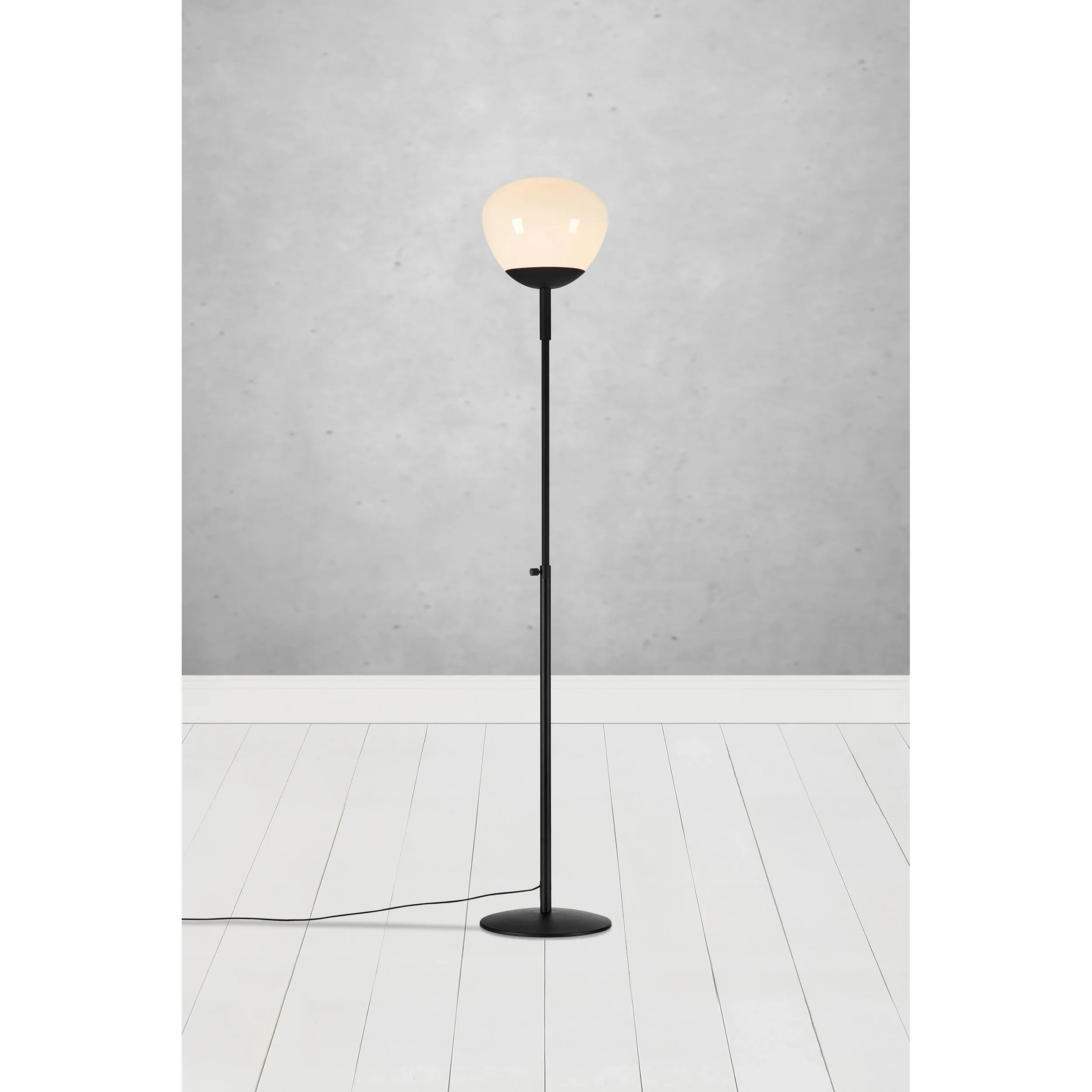 Lampa podłogowa Rise 1-punktowa w kolorze czarnym z białym kloszem, stojąca na okrągłej, czarnej podstawie. Ujęcie frontalne, w tle szare ściany i biała podłoga. Kabel zasilający widoczny obok nogi lampy.