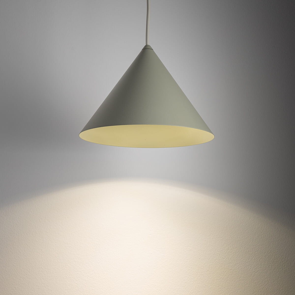 Lampa wisząca Zenith S w kolorze szałwii, o kształcie stożkowym, zawieszona na białym przewodzie. Emituje ciepłe światło, rzucając delikatny cień na ścianę. Wewnątrz miejsce na żarówkę GU10. Tło w jasnym odcieniu szarości.