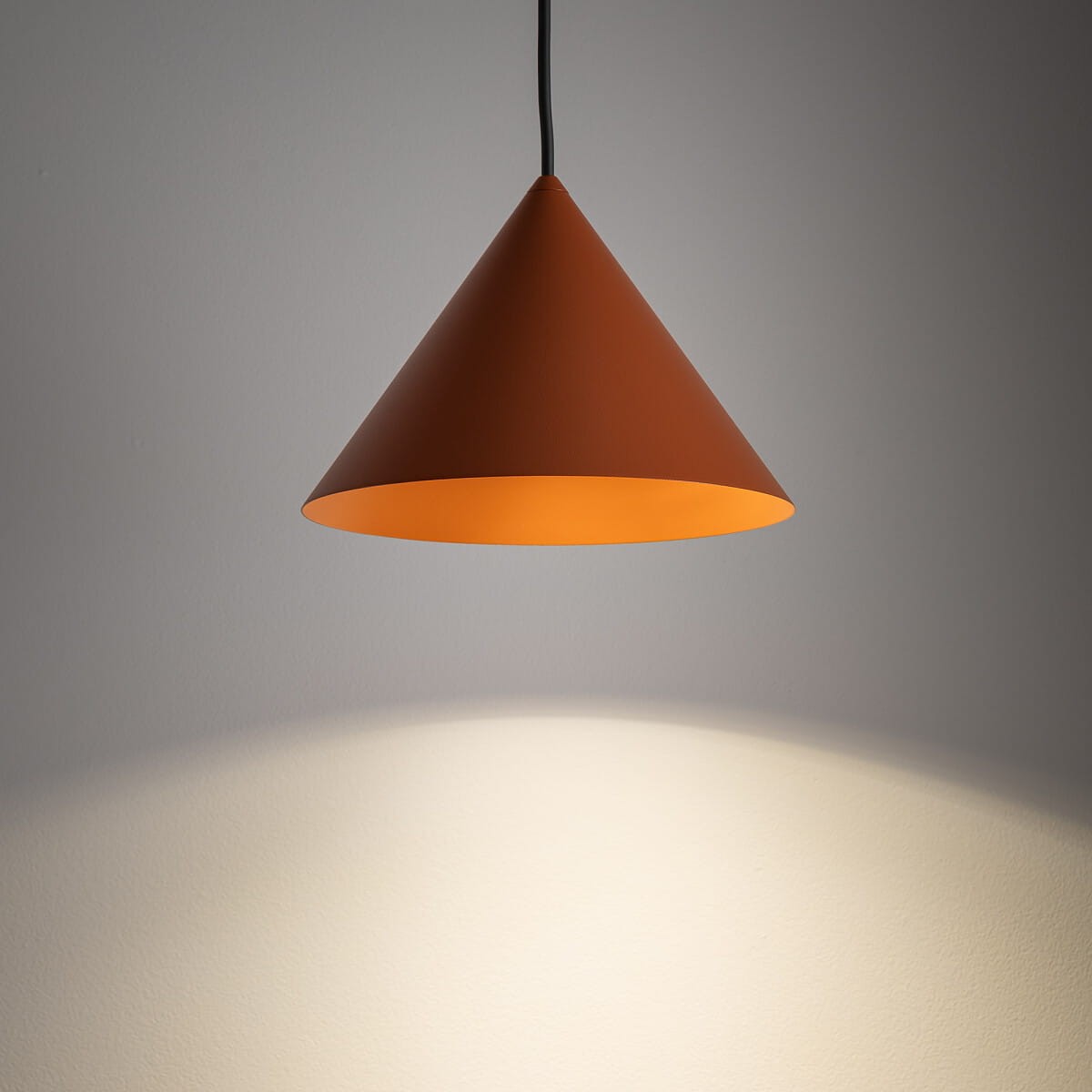 Lampa wisząca Zenith S w ceglastym kolorze, o kształcie stożka, z jedną oprawą GU10. Zawieszona na czarnym przewodzie, emitująca ciepłe światło, z widocznym cieniem na ścianie. Idealna do nowoczesnych wnętrz.