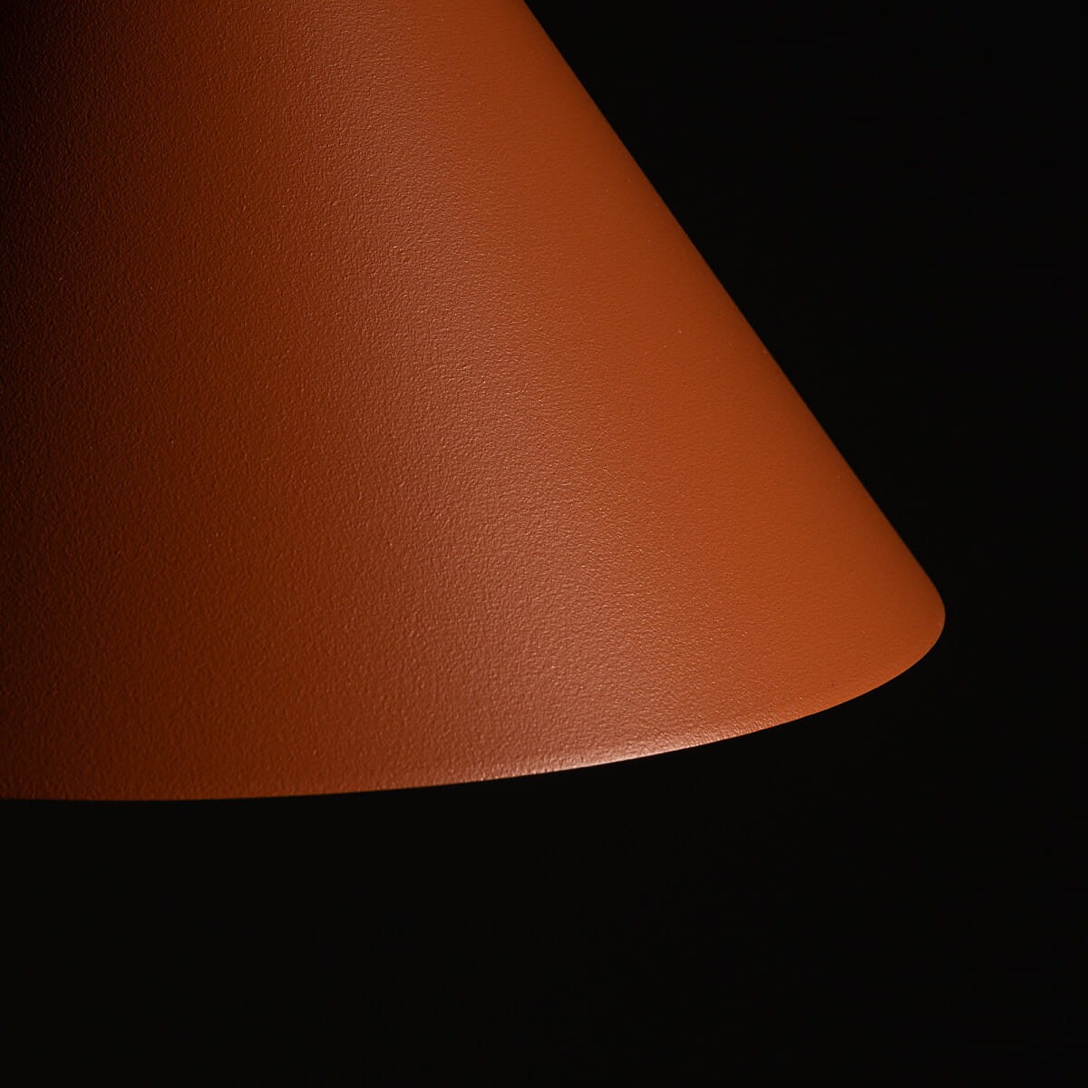 Lampa wisząca Zenith S w kolorze ceglastym, z kloszem w kształcie stożka. Ujęcie z boku na teksturowaną powierzchnię. Produkt zawiera miejsce na jedną żarówkę GU10, idealny do nowoczesnych wnętrz. Tło ciemne, podkreślające kolor lampy.