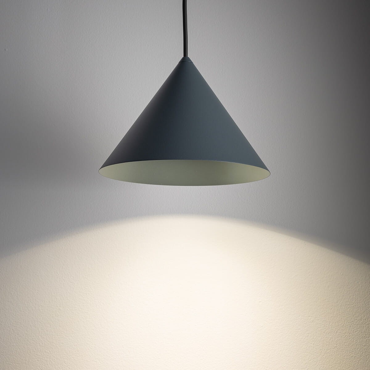 Lampa wisząca Zenith S w kształcie stożka, w kolorze niebieskim, z jednym gniazdem GU10. Zawieszona na czarnym przewodzie, emitująca światło na białą ścianę. Prosta, nowoczesna forma bez dodatkowych zdobień.