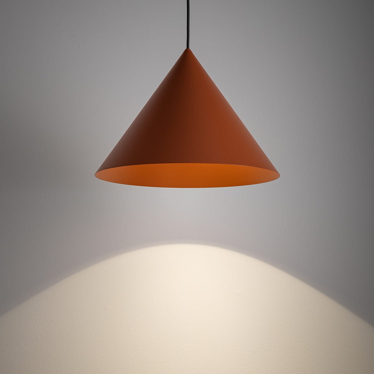 Lampa wisząca Zenith M w kolorze ceglastym, w kształcie stożka, zawieszona na czarnym kablu. Emituje światło w kierunku ściany, tworząc subtelny cień. Idealna do nowoczesnych wnętrz.