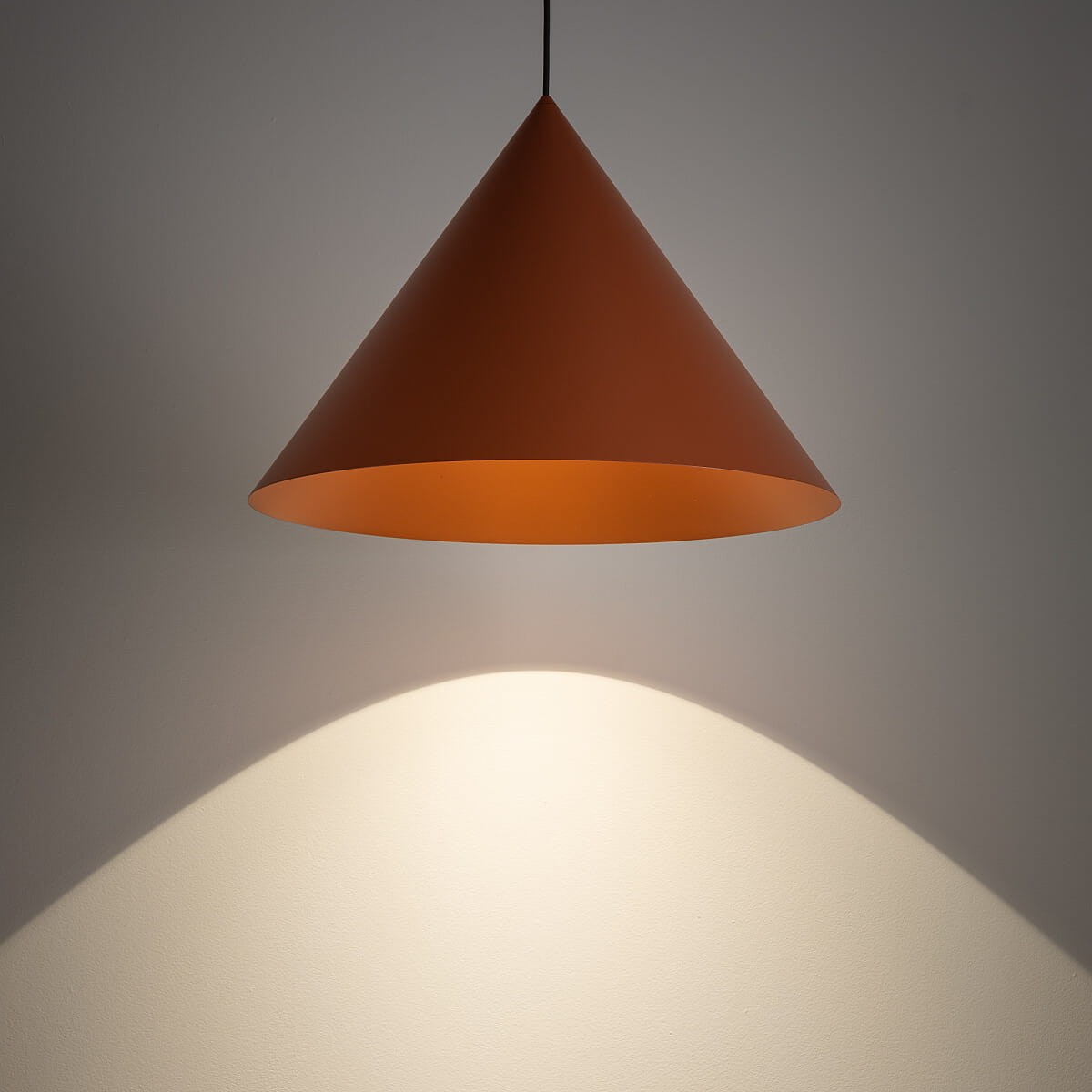 Lampa wisząca Zenith L w kształcie stożka, w kolorze ceglastym, z pojedynczym gniazdem GU10. Ujęcie z dołu pokazuje cień na ścianie, podkreślając jej minimalistyczny design. Mosiężny klosz zawieszony na czarnym kablu.