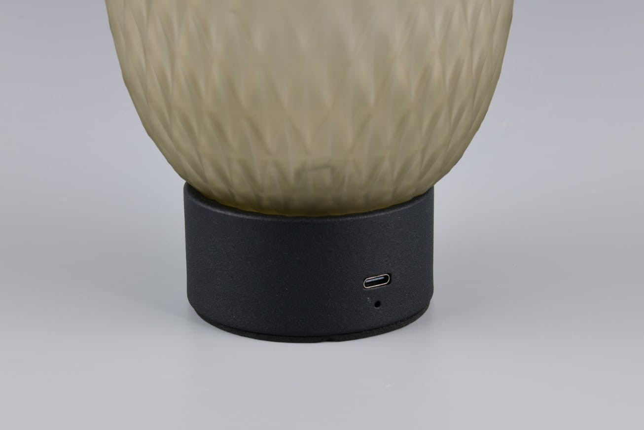 Lampa stołowa Facette z przezroczystym kloszem w kształcie owalnym, umieszczona na czarnej podstawie. Widoczny port USB-C z boku podstawy. Klosz ma delikatną, teksturowaną powierzchnię.