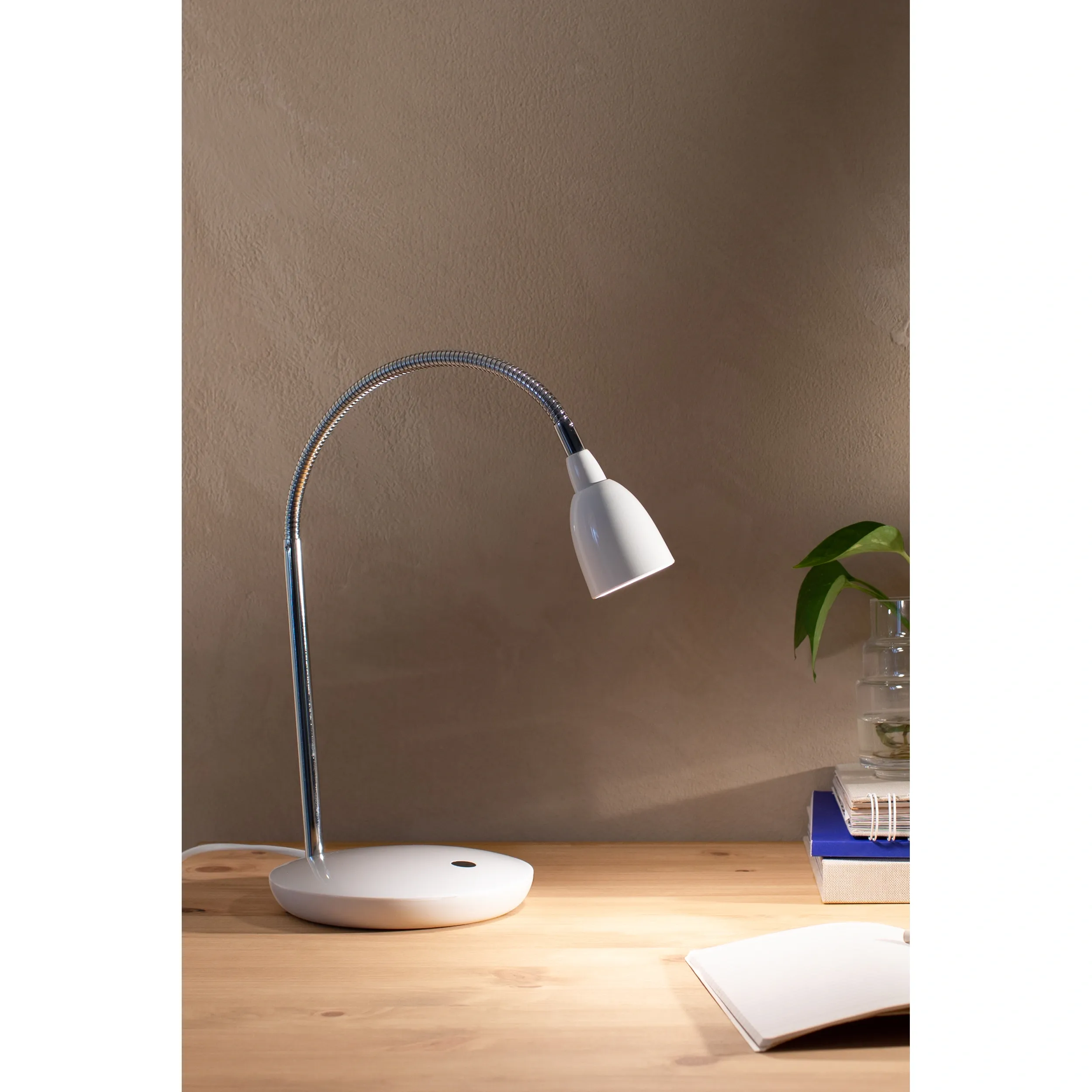 Biała lampa biurkowa Tulip LED z elastycznym ramieniem, stojąca na okrągłej podstawie. Klosz o zaokrąglonym kształcie skierowany w dół, w tle widoczne książki oraz roślina w doniczce. Ujęcie z boku na drewnianym biurku.