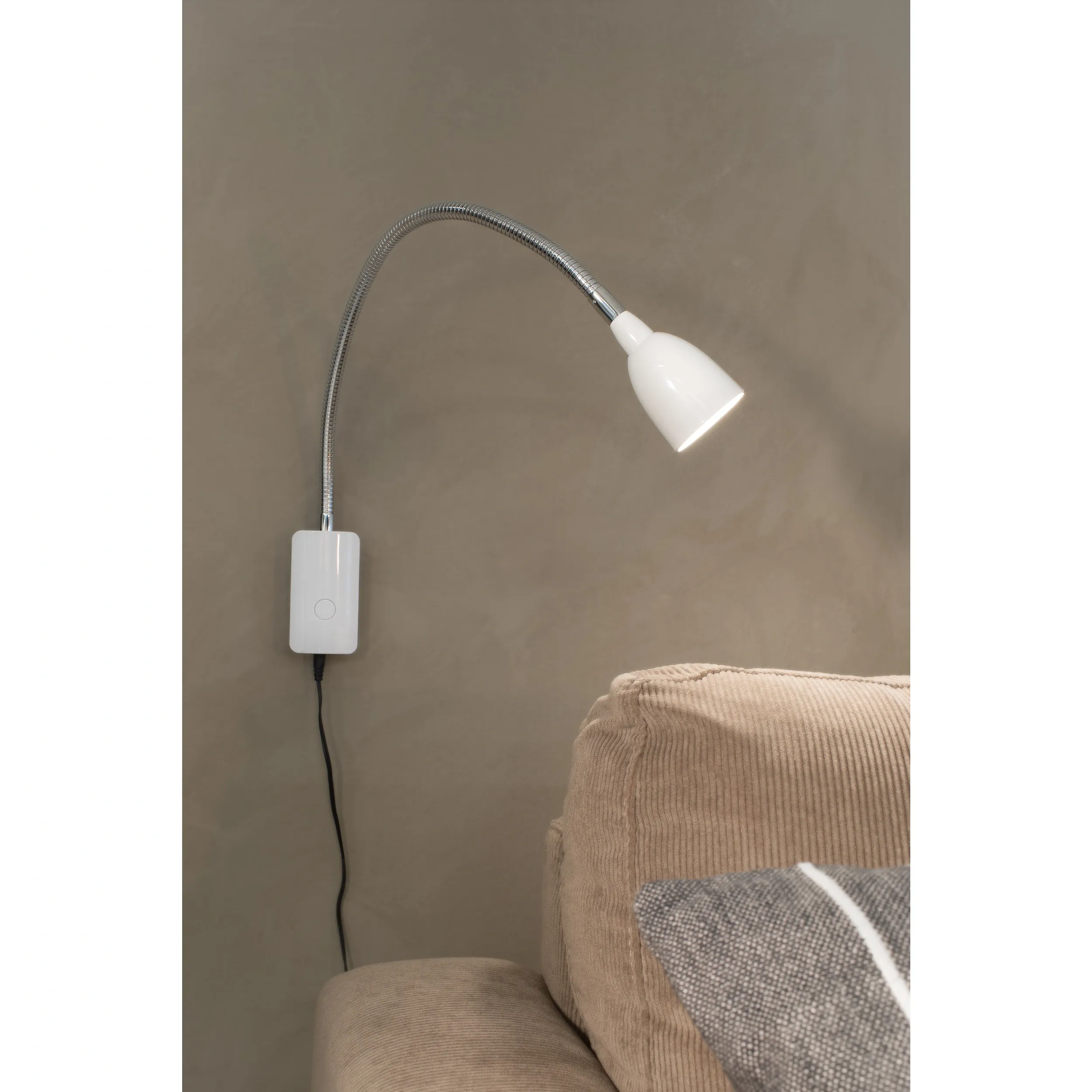 Tulip LED kinkiet 1-punktowy w białej obudowie, z elastycznym metalowym ramieniem, zamontowany na ścianie obok kanapy. Widoczny włącznik na ścianie oraz przewód zasilający. W głowicy lampy znajduje się źródło światła.