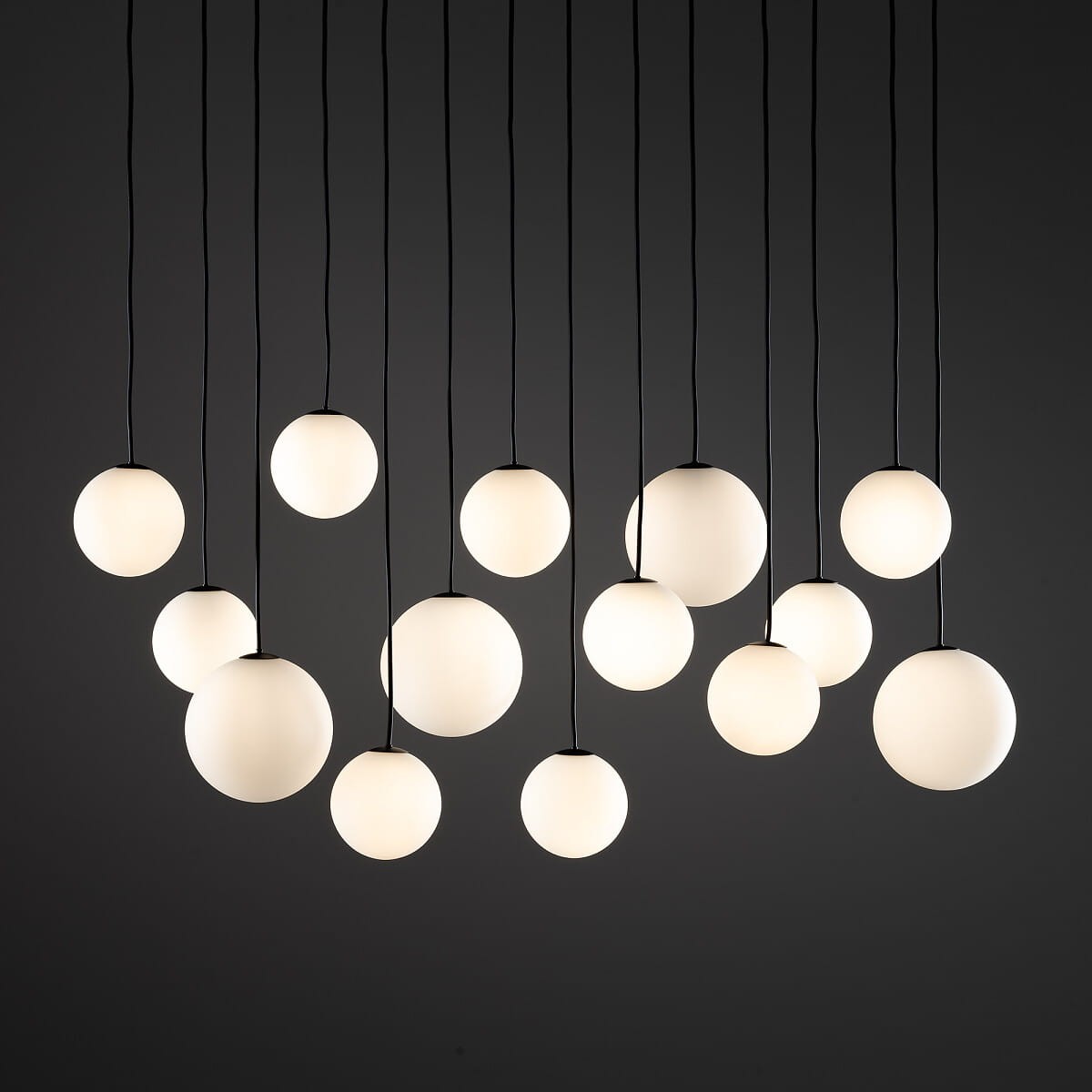 Lampa wisząca Snowball XIV z 14 źródłami światła G9, w czarnej oprawie. Okrągłe, białe klosze o matowej powierzchni zawieszone na cienkich, czarnych przewodach. Ułożone w asymetrycznym wzorze, na ciemnym tle. Idealna do nowoczesnych wnętrz.