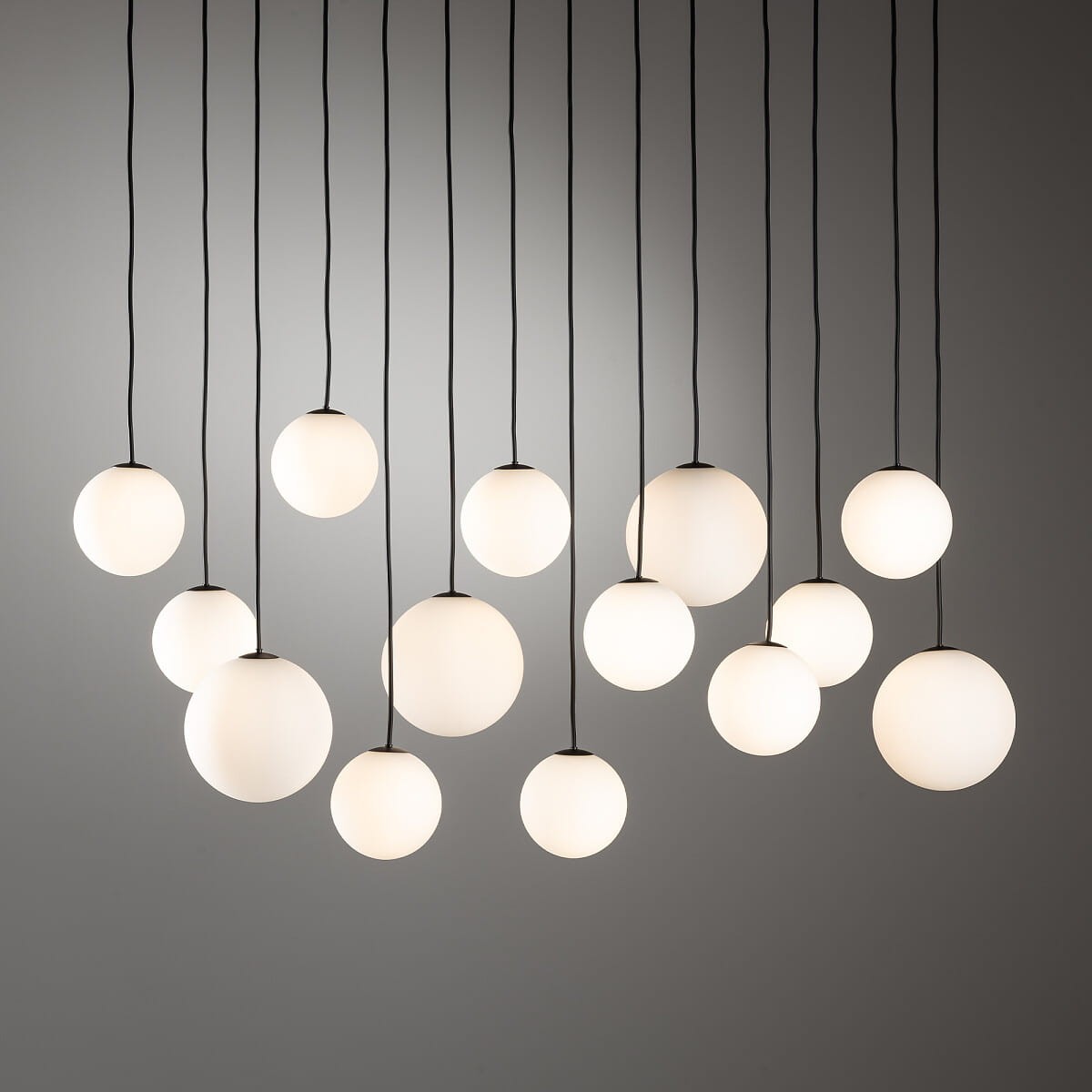 Lampa wisząca Snowball XIV z 14 kulistymi kloszami w białym kolorze, zawieszonymi na czarnych linkach. Klosze o matowej powierzchni różnej wielkości, wiszące w różnych wysokościach na prostym tle.