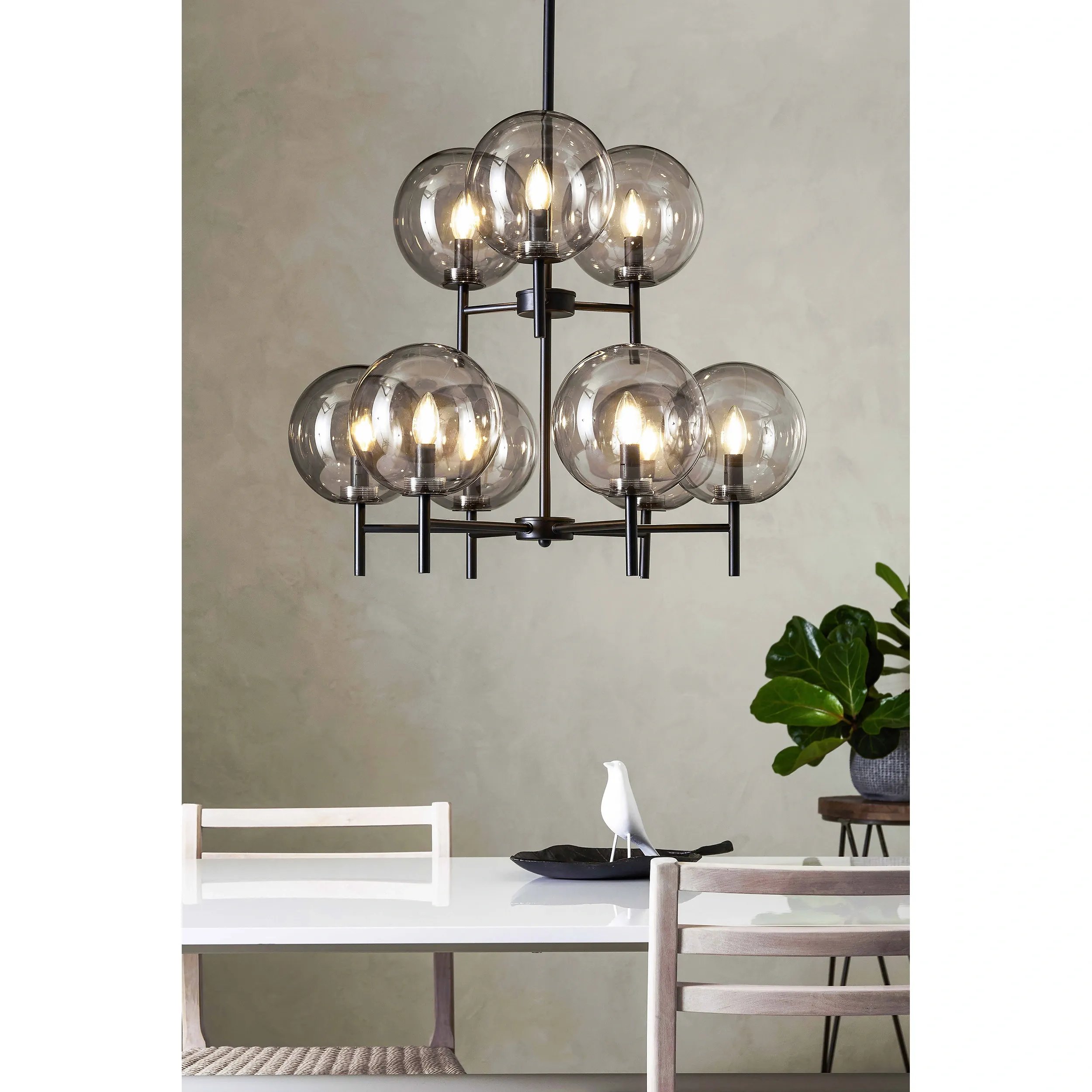 Lampa wisząca Crown z 9 punktami świetlnymi, w kolorze czarnym z przydymionymi kloszami. Ujęcie z góry, zawieszona nad stołem w jadalni. W tle widoczna roślina i biały stół z drewnianymi krzesłami.