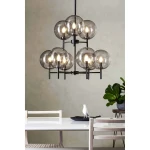 Lampa wisząca Crown z 9 punktami świetlnymi, w kolorze czarnym z przydymionymi kloszami. Ujęcie z góry, zawieszona nad stołem w jadalni. W tle widoczna roślina i biały stół z drewnianymi krzesłami.