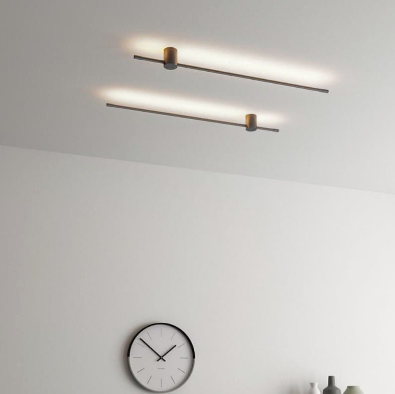 Kinkiet Catrina 120 w kolorze czarnym z jedną diodą LED o mocy 17W i temperaturze barwowej 4000K. Zamontowany na suficie, z minimalistycznym, prostokątnym kształtem oraz subtelnym oświetleniem. W tle widoczny zegar ścienny.