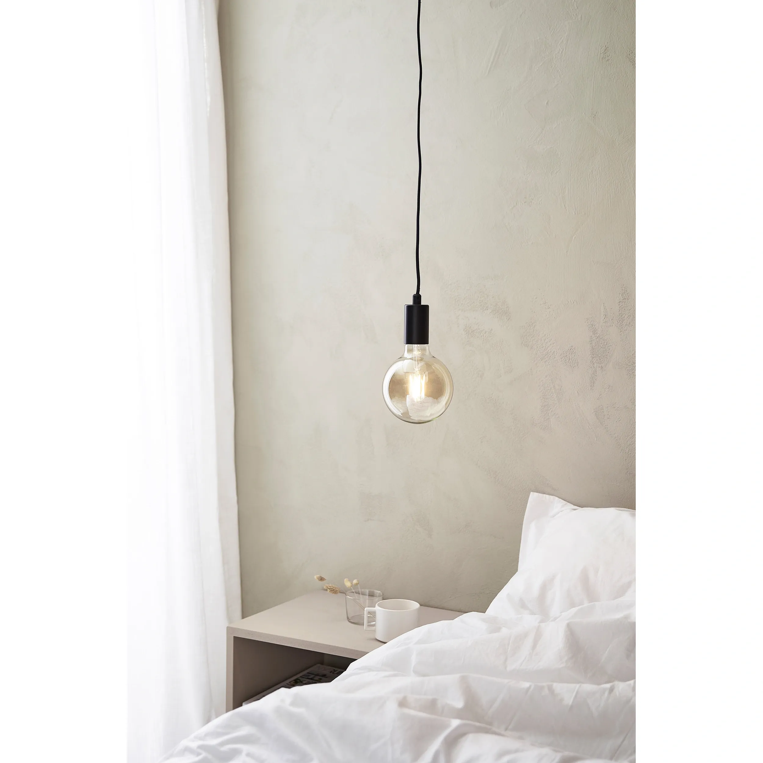 Lampa wisząca Sky 1-punktowa w kolorze czarnym, z przezroczystą żarówką, zawieszona na długim kablu. Umieszczona nad stolikiem nocnym, w jasnym wnętrzu z szarymi ścianami i białą pościelą.