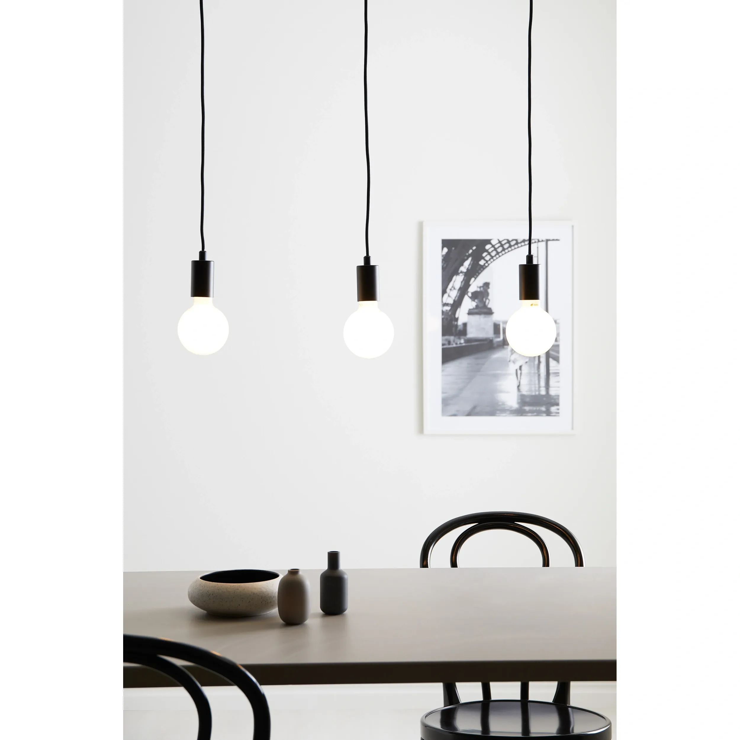 Lampa wisząca Sky 3-punktowa w kolorze czarnym, zawieszona na długich kablach. Trzy okrągłe żarówki w metalowych oprawach, na tle jasnej ściany. W dolnej części widoczny stół z akcesoriami, w tym ceramiczną misą i dwoma wazonami.