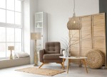 Lampa wisząca Janne w naturalnym kolorze, z kloszem w formie plecionki, zawieszona nad białym stolikiem. W tle brązowy fotel, dywan z juty oraz półka z dekoracjami. Jasne, minimalistyczne wnętrze.