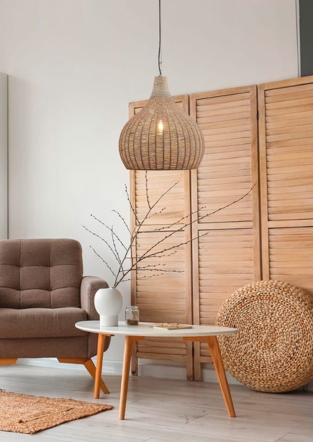 Lampa wisząca Janne z kloszem w naturalnym kolorze, wykonana z plecionego materiału, zawieszona na cienkim przewodzie. Produkt umieszczony w aranżacji wnętrza z brązowym fotelem i stolikiem.