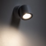 Lampa sufitowa Oscar w kolorze białym, z reflektorem 1xGU10, montowana na ścianie. Widać jasne światło emitowane przez żarówkę oraz cień na ścianie. Kształt okrągły, prosty design, idealna do oświetlenia wnętrz. IP54, odporna na wilgoć.