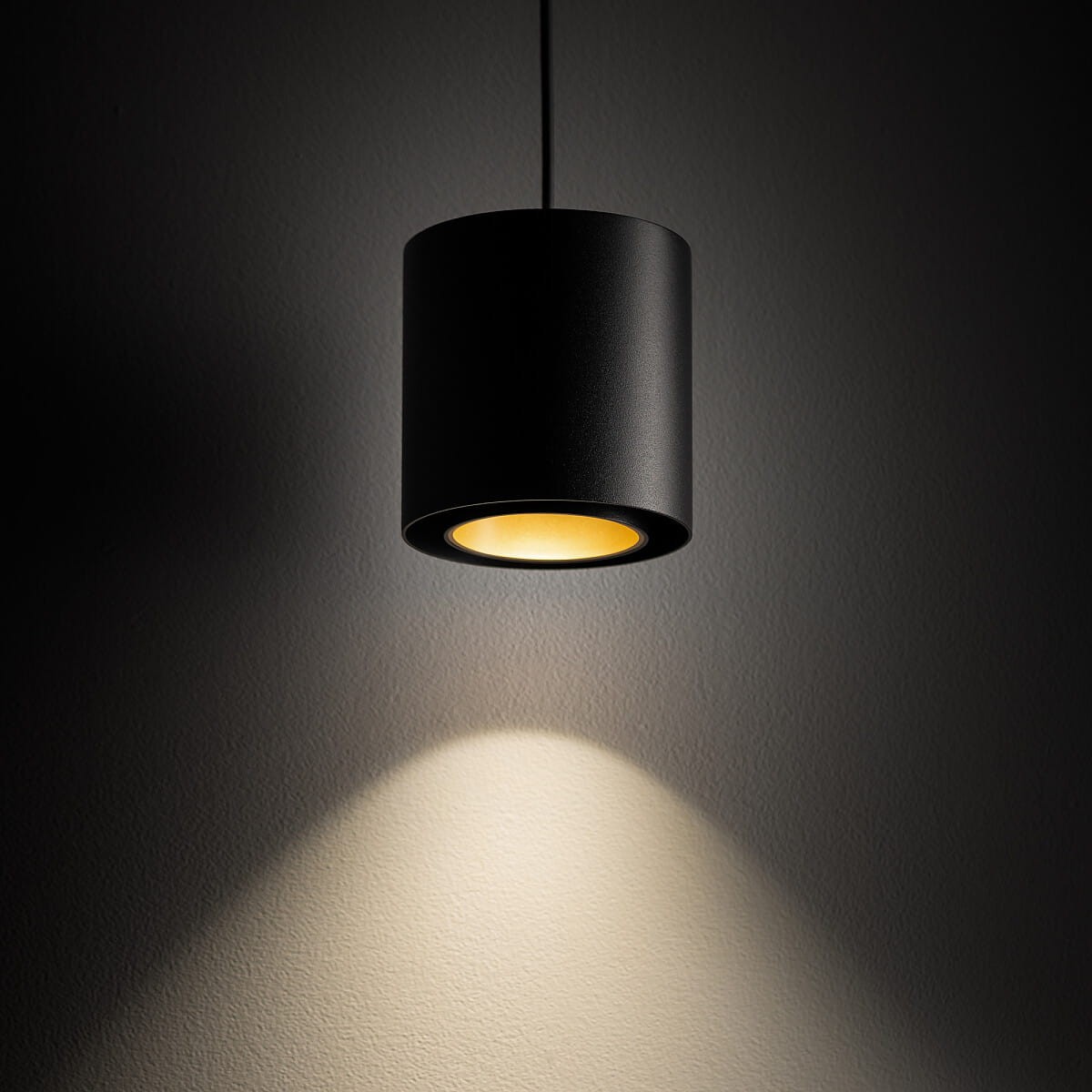 Lampa wisząca Point Duo w kolorze czarnym z złotym wnętrzem, o cylindrycznym kształcie. Zawieszona na cienkim kabelku, emituje ciepłe światło, które rzuca cień na ścianę. Idealna do nowoczesnych wnętrz.