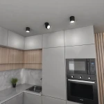 Czarna lampa sufitowa Arto z pojedynczym spotem GX53, zamontowana na suficie w nowoczesnej kuchni. Otoczenie obejmuje szare i drewniane szafki, a w tle widoczny jest zlewozmywak i sprzęt AGD. Oświetlenie rozproszone, minimalistyczny design.