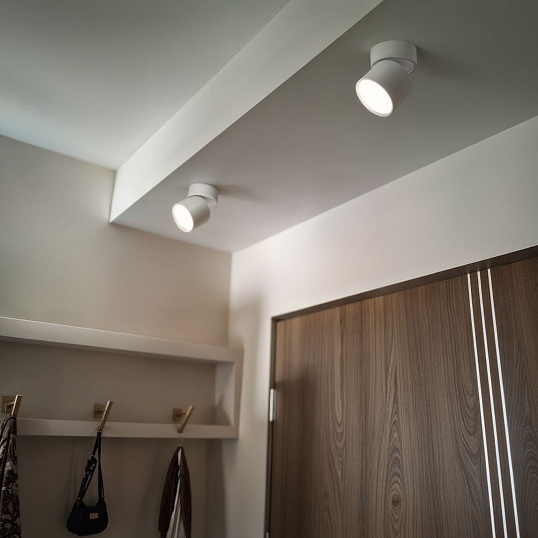 Biała lampa sufitowa Arto w formie spotu z jednym źródłem światła GX53, zamontowana na suficie, skierowana w dół. W tle widoczna drewniana drzwi i półka z wieszakami. Ściany w jasnym kolorze.