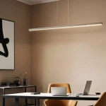 Lampa wisząca Linia 120 z białym wykończeniem, o długości 1 metra, zawieszona na dwóch cienkich linkach. Emituje światło LED o mocy 15W. Wnętrze biura z jasną ścianą, drewnianymi krzesłami i stołem z laptopem.
