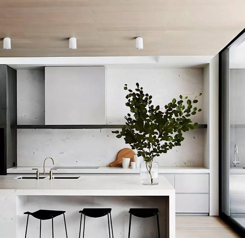 Biała lampa sufitowa tuba kierunkowa, zamontowana na drewnianym suficie, o minimalistycznym designie. W tle nowoczesna kuchnia z białymi szafkami i czarnymi krzesłami, z widocznym blatem i rośliną w szklanej wazonie.