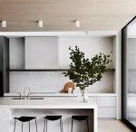 Biała lampa sufitowa tuba kierunkowa, zamontowana na drewnianym suficie, o minimalistycznym designie. W tle nowoczesna kuchnia z białymi szafkami i czarnymi krzesłami, z widocznym blatem i rośliną w szklanej wazonie.