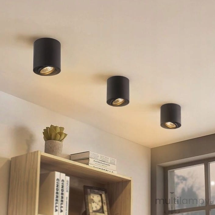 Mini lampa sufitowa tuba w kolorze czarnym, zamontowana na suficie. Trzy lampy w równych odstępach, skierowane w dół. W tle widoczne półki z książkami i rośliną. Proste, nowoczesne wzornictwo.