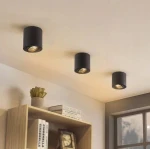 Mini lampa sufitowa tuba w kolorze czarnym, zamontowana na suficie. Trzy lampy w równych odstępach, skierowane w dół. W tle widoczne półki z książkami i rośliną. Proste, nowoczesne wzornictwo.