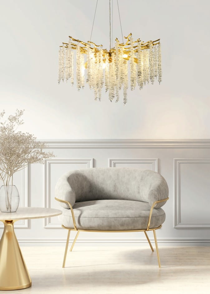 Lampa wisząca Dijon 10xG9 w złotym kolorze, z dekoracyjnymi, przezroczystymi kryształkami zwisającymi, zawieszona na cienkim przewodzie. Produkt umieszczony w jasnym wnętrzu, na tle białej ściany, obok szarego fotela i złotego stolika.
