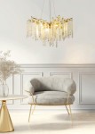 Lampa wisząca Dijon 10xG9 w złotym kolorze, z dekoracyjnymi, przezroczystymi kryształkami zwisającymi, zawieszona na cienkim przewodzie. Produkt umieszczony w jasnym wnętrzu, na tle białej ściany, obok szarego fotela i złotego stolika.