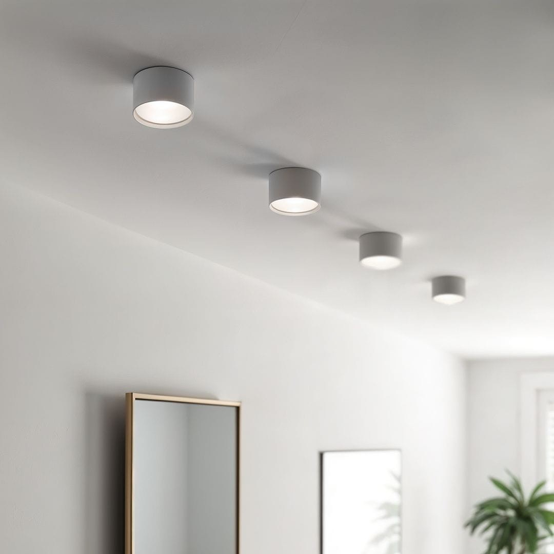 Biała lampa sufitowa tuba Leto z jednym gniazdem GX53, zamontowana na suficie. Widoczne cztery lampy w równych odstępach, emitujące jasne światło. W tle lustro w złotej ramie oraz roślina doniczkowa.