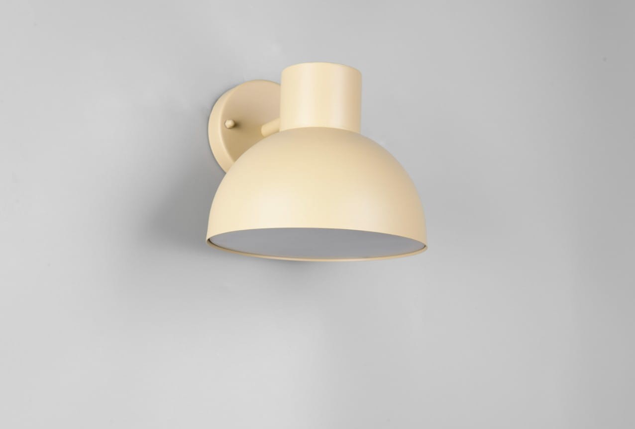 Kinkiet zewnętrzny Elbe w kolorze piaskowym, z okrągłą, matową osłoną i prostym uchwytem. Zamontowany na ścianie, prezentuje minimalistyczny design. Wewnątrz miejsce na żarówkę E27. Tło zdjęcia szare, co podkreśla kolor lampy.