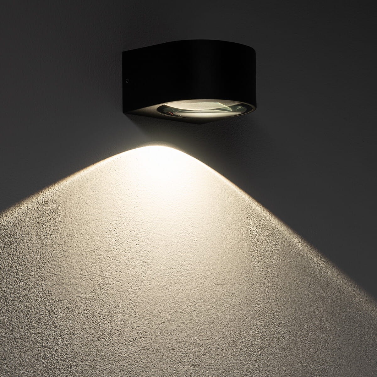 Kinkiet elewacyjny Nel w kolorze czarnym z jedną diodą LED (5W, 3000K) zamontowany na ścianie. Oświetlenie podkreśla teksturę powierzchni, emitując ciepłe światło w kształcie łuku. Produkt ma nowoczesny, minimalistyczny design.
